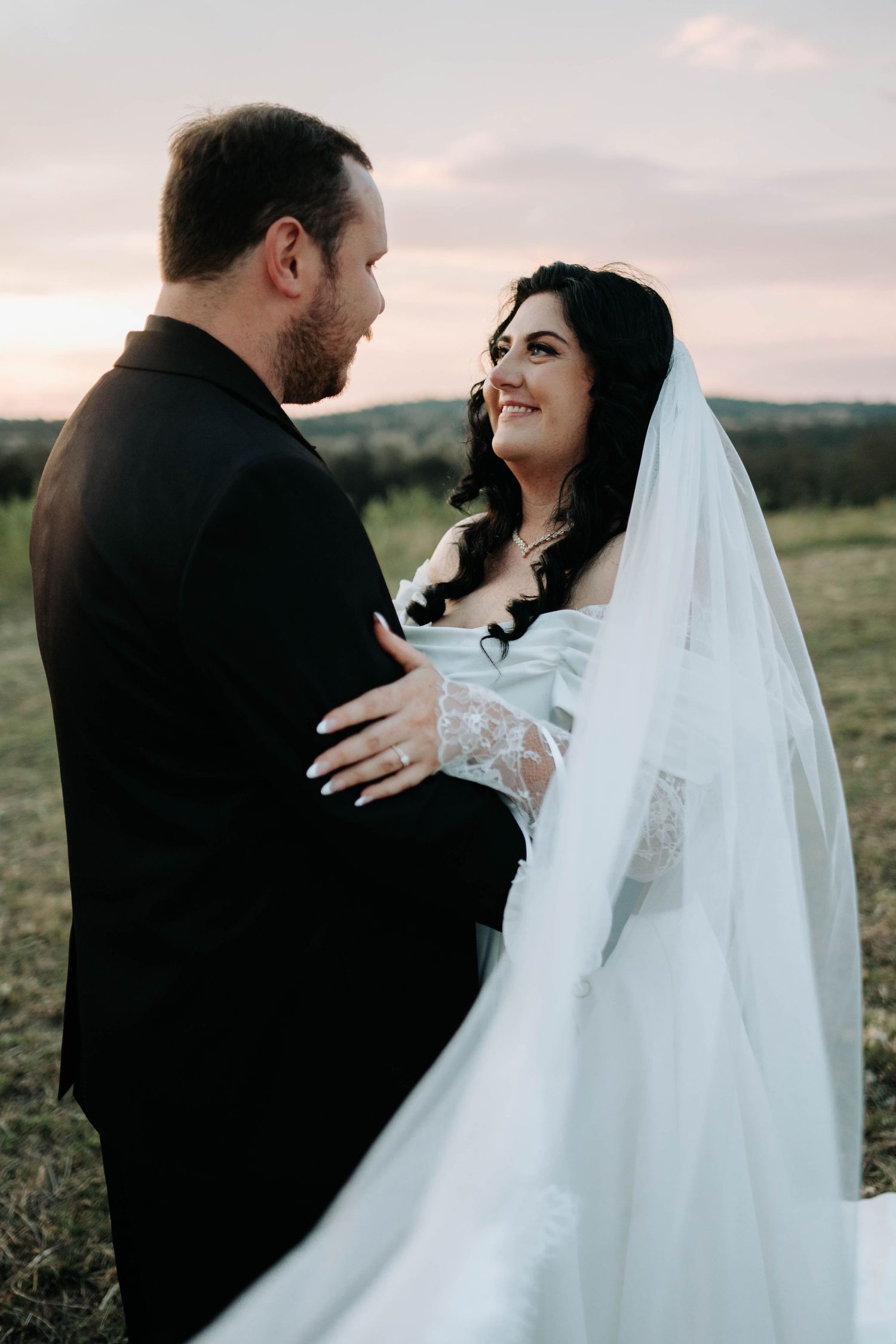 Romantic wedding couple embraces during sunset photoshoot in countryside field with flowing white veil.