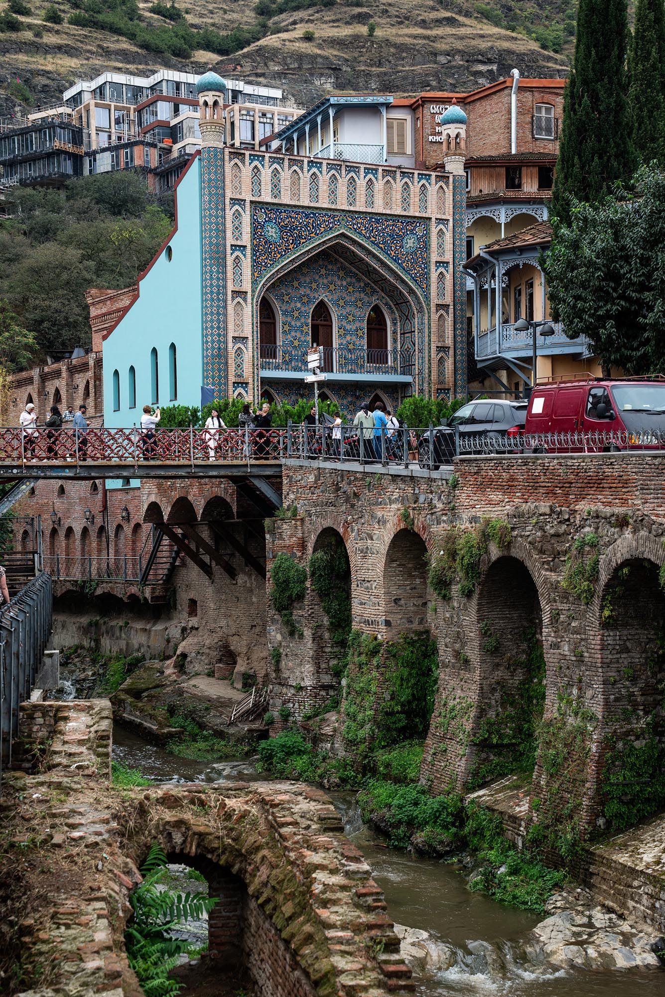 Historic stone bridge with arches spans a river gorge in Tbilisi, Georgia with traditional architecture visible above.