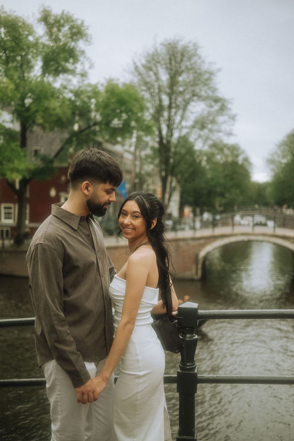 Couple holding hands on a bridge over an Amsterdam canal, woman in white dress smiling, lush green trees in background.