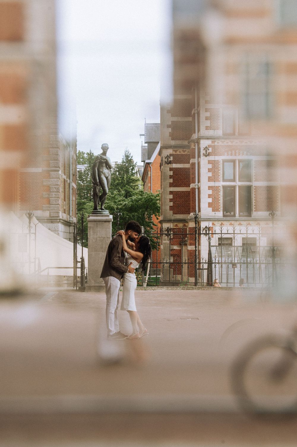 Couple embracing romantically in a European city square with historic brick buildings and a statue in the background.