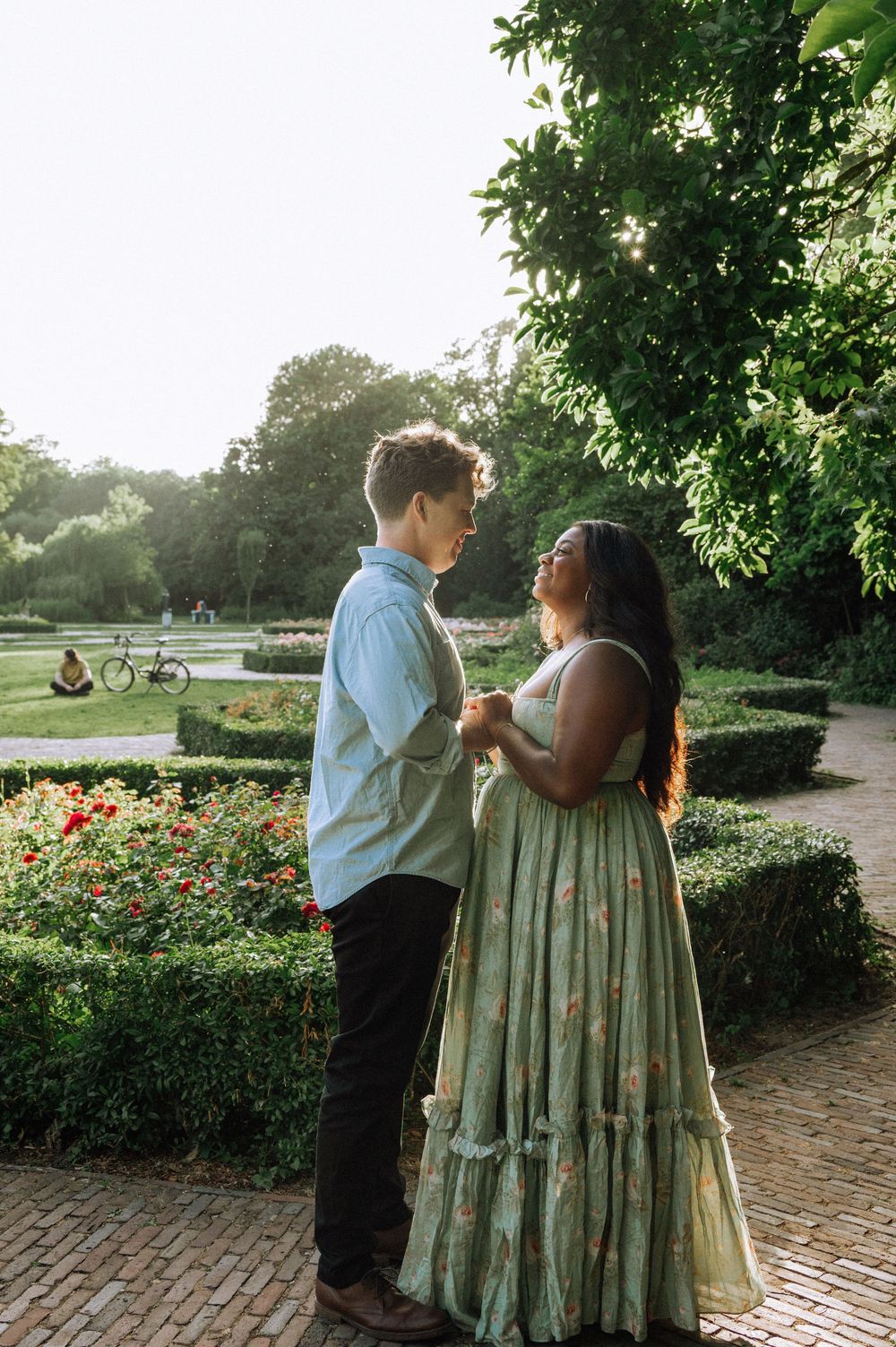 Couple holding hands in a lush garden park at golden hour, woman in floral green dress, man in blue shirt.