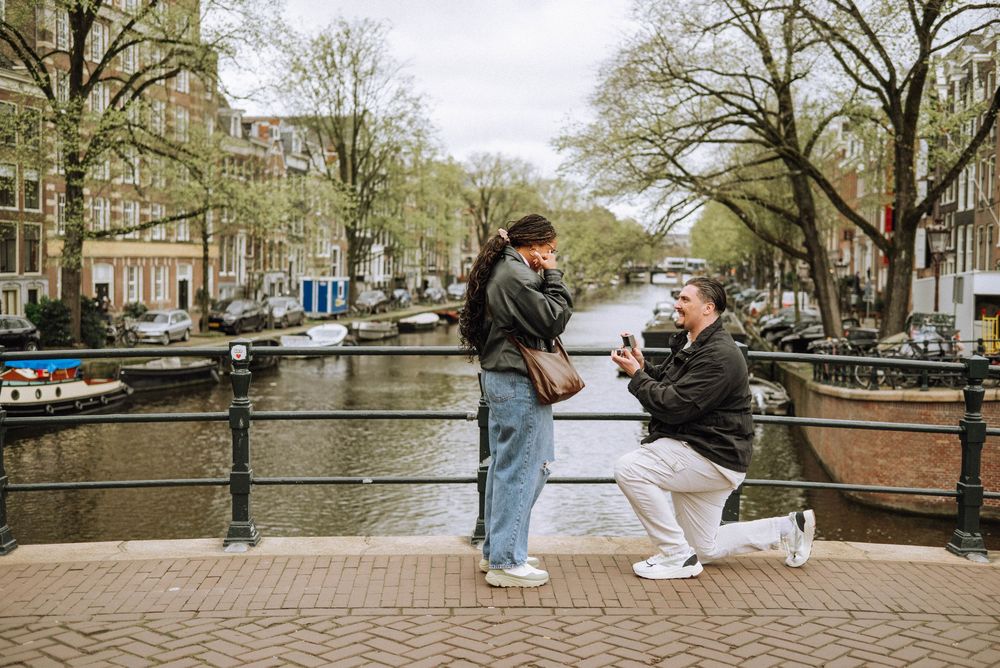 Man proposes to woman on a bridge over an Amsterdam canal, surrounded by trees and historic Dutch buildings.