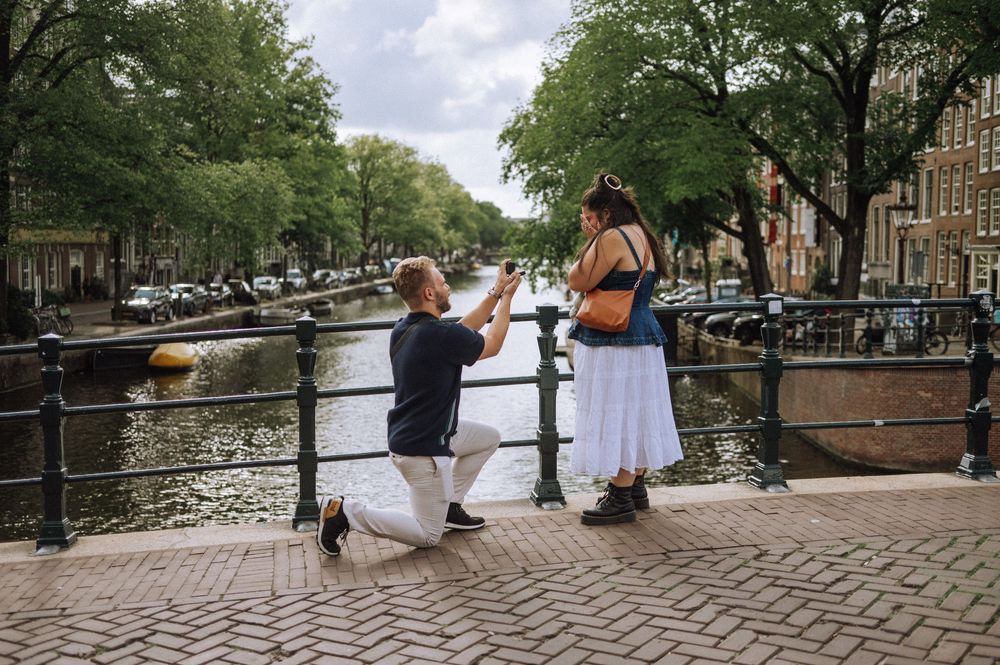 People relaxing along a scenic canal in Amsterdam with trees and historic buildings.