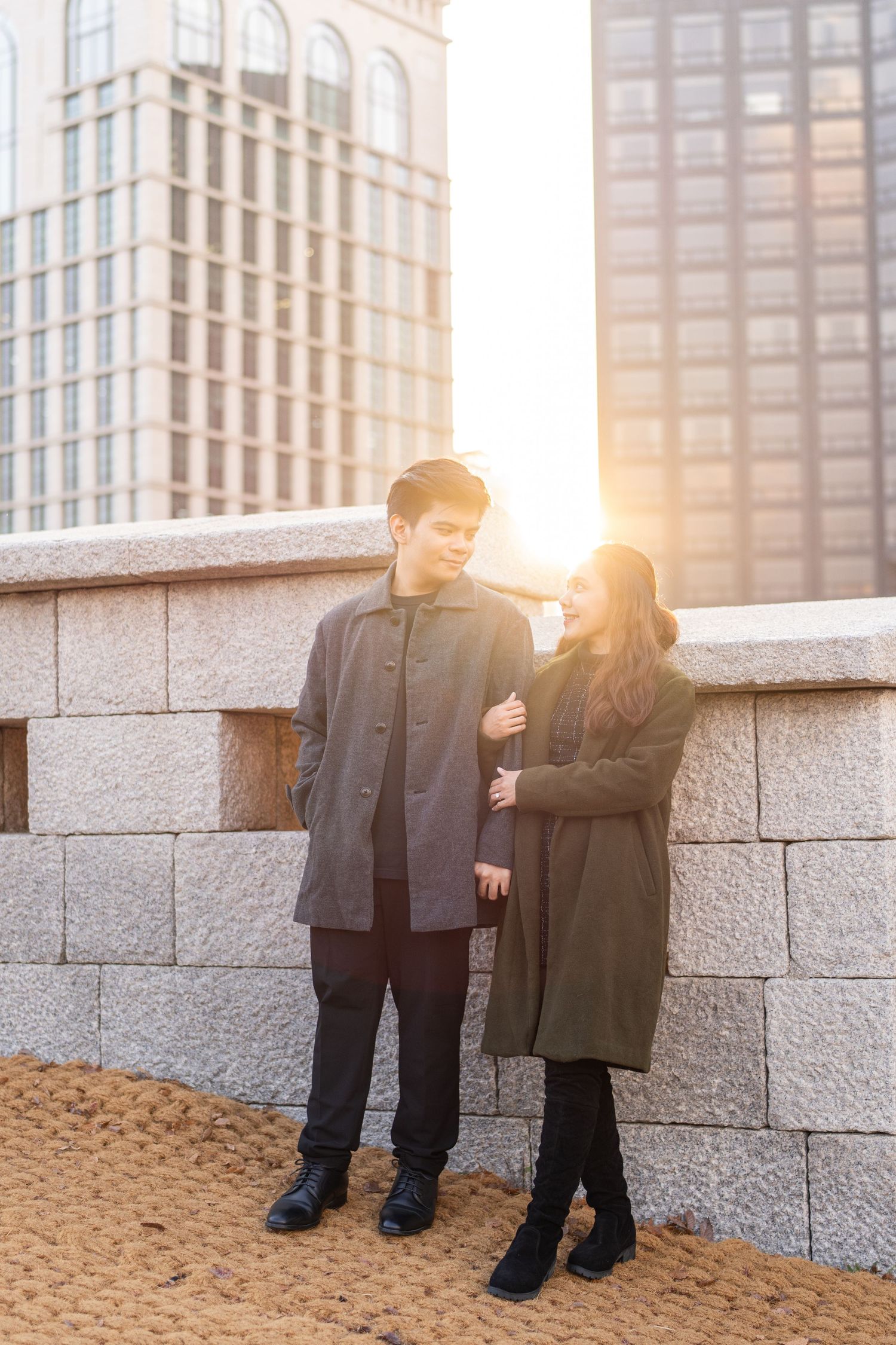 Two people in winter coats stand against a stone wall as golden sunlight creates a warm glow in an urban setting.