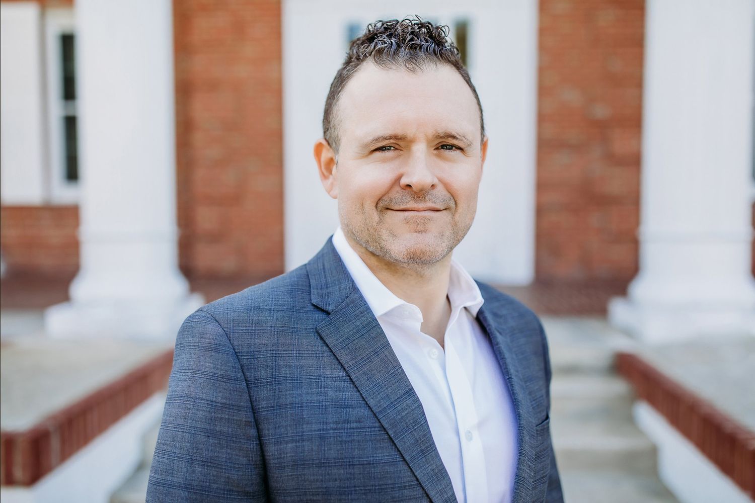 Professional headshot portrait of someone in a navy suit jacket and white dress shirt against a brick building exterior.