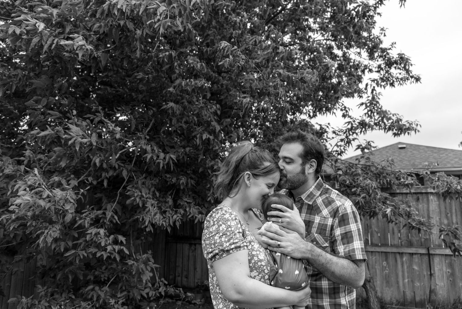 B/W family photo with 2 new parents. The baby's facing away from the camera with mom kissing her head and dad kissing moms forehead