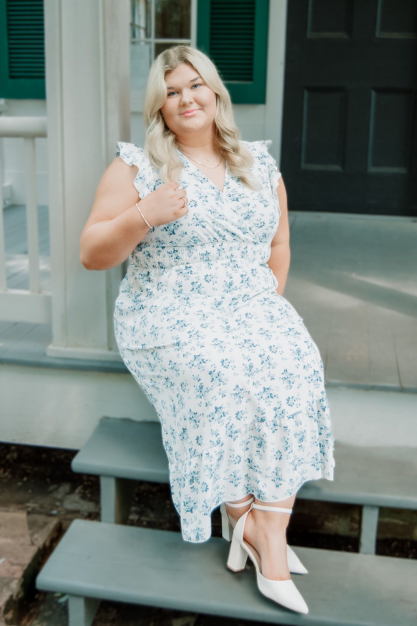 Woman in a floral print dress and white heels poses on outdoor steps.