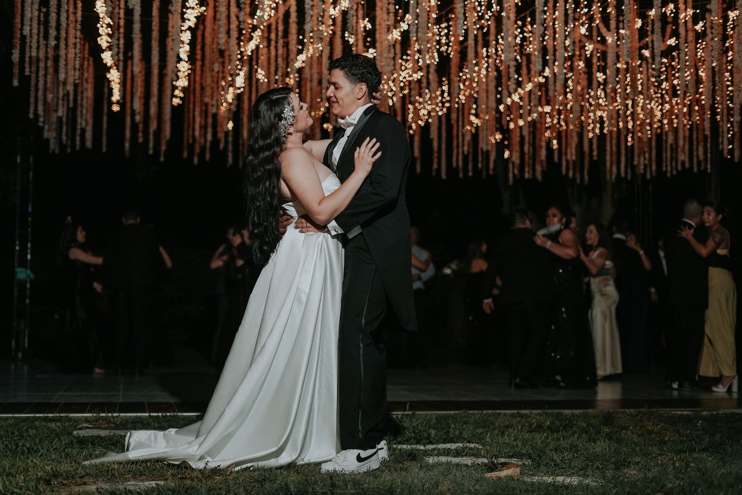 A couple embraces in front of a glowing backdrop of warm string lights at a wedding reception.