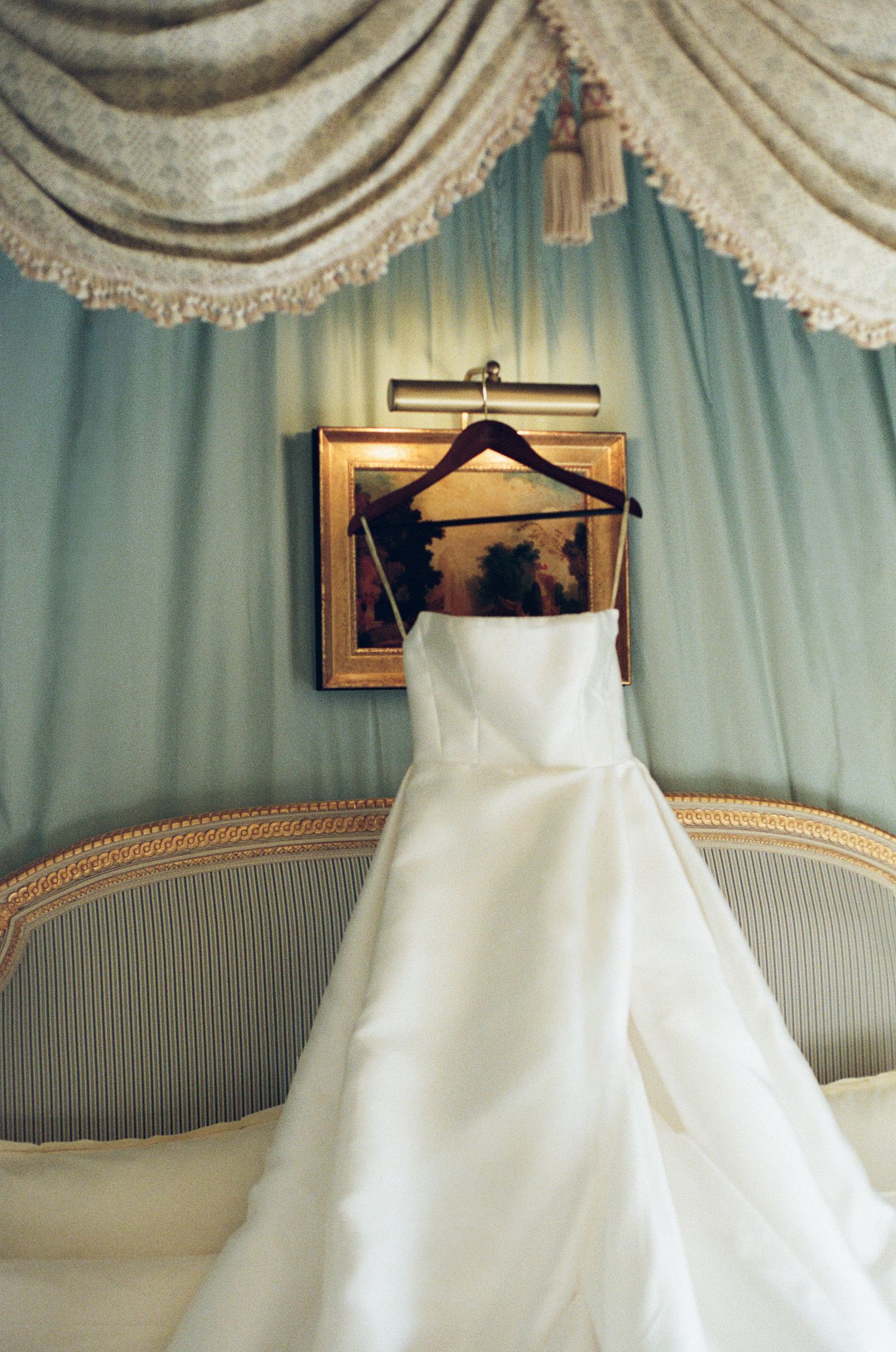 A series of elegant white wedding dresses displayed on hangers against a blue-green wall with ornate molding and drapes.