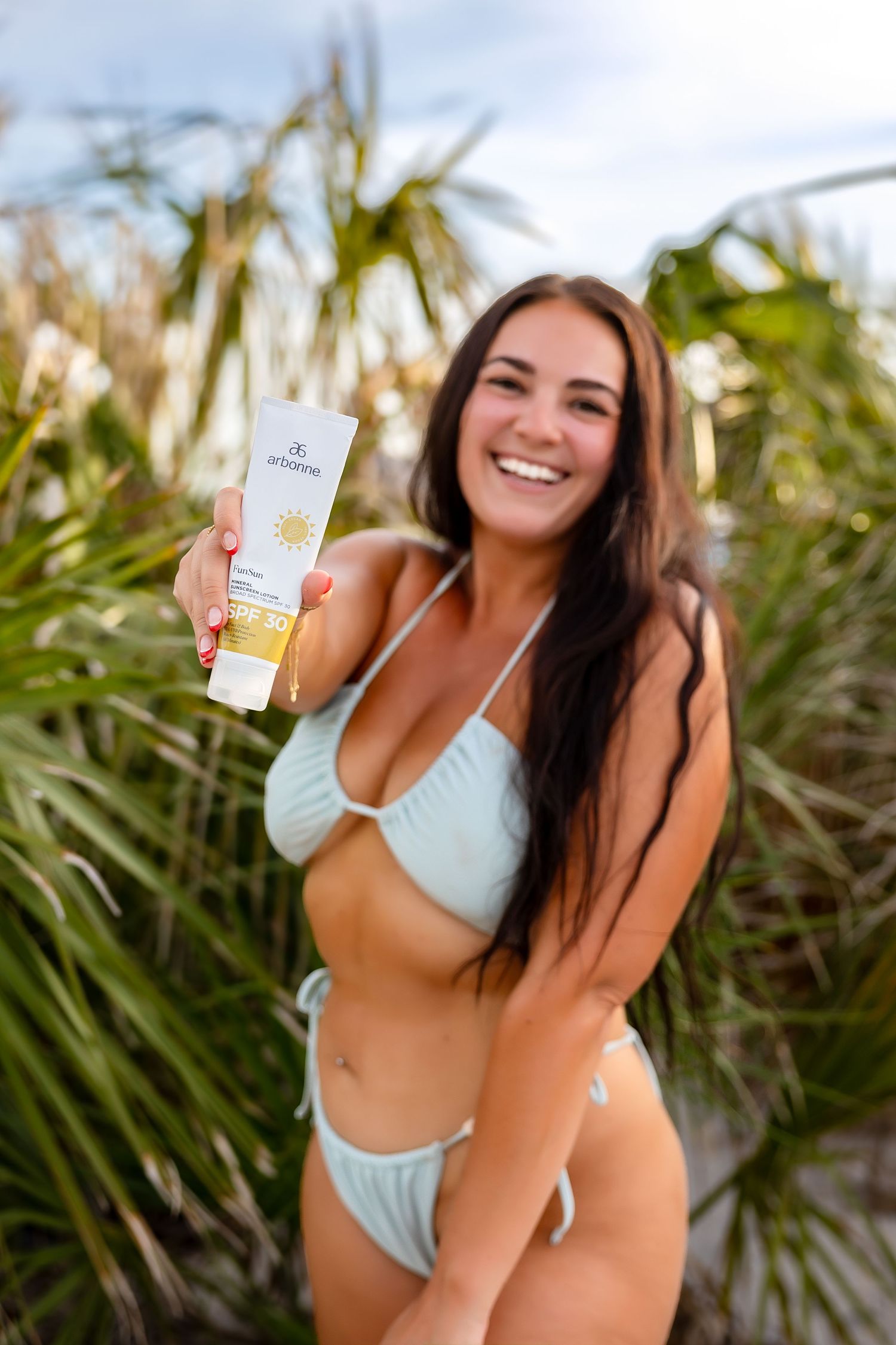 Smiling woman in light blue bikini holding Arbonne sunscreen product outdoors.