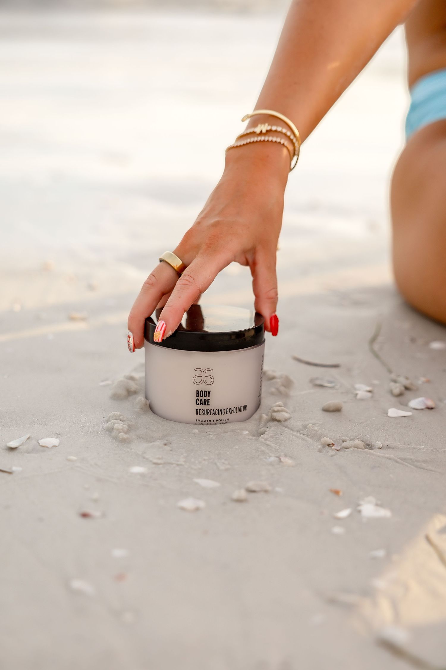 Woman in blue bikini reaching for Arbonne body care rejuvenating cream on sand.