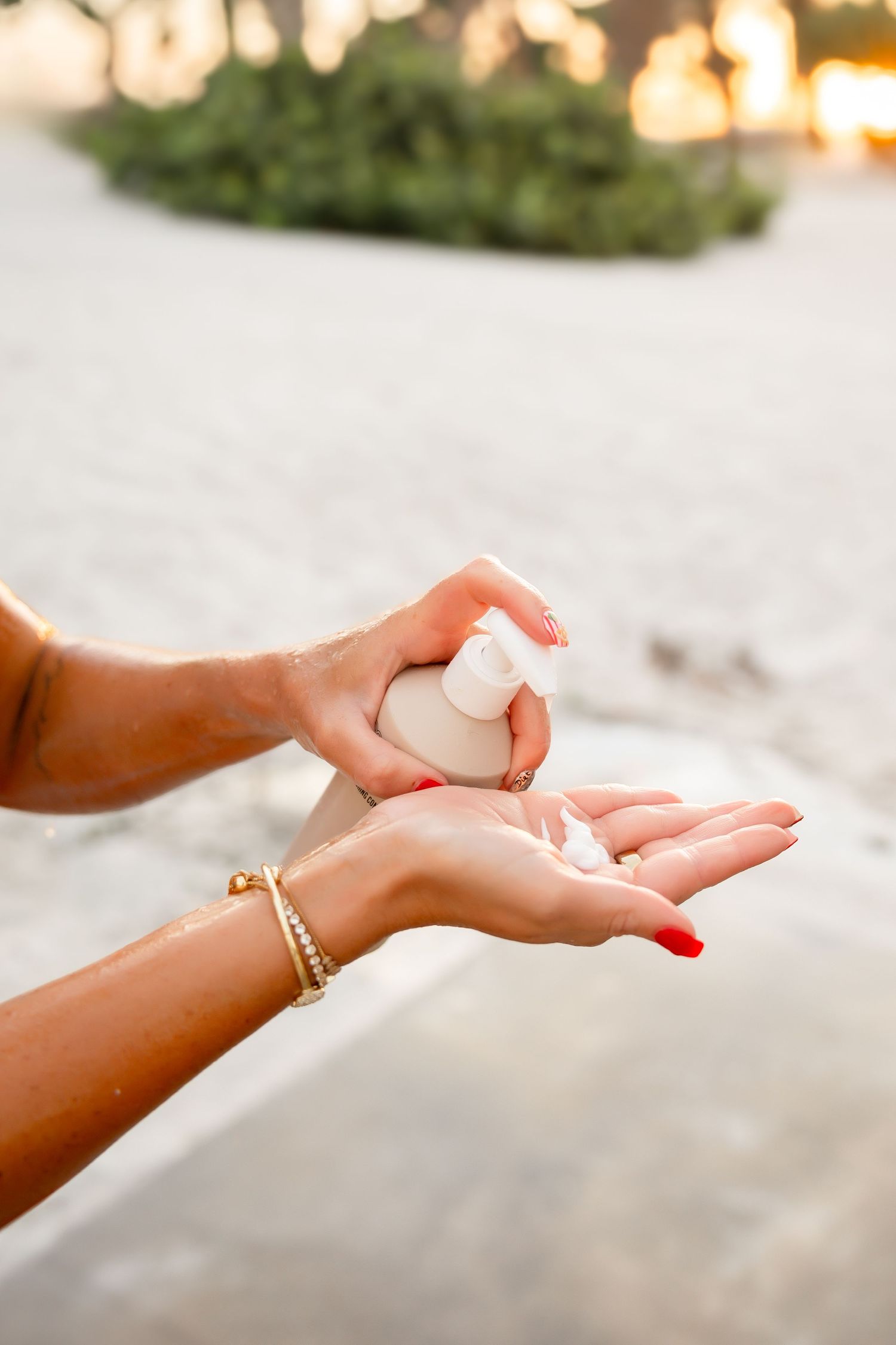 Hands dispensing lotion from white pump bottle on sandy beach at sunset.