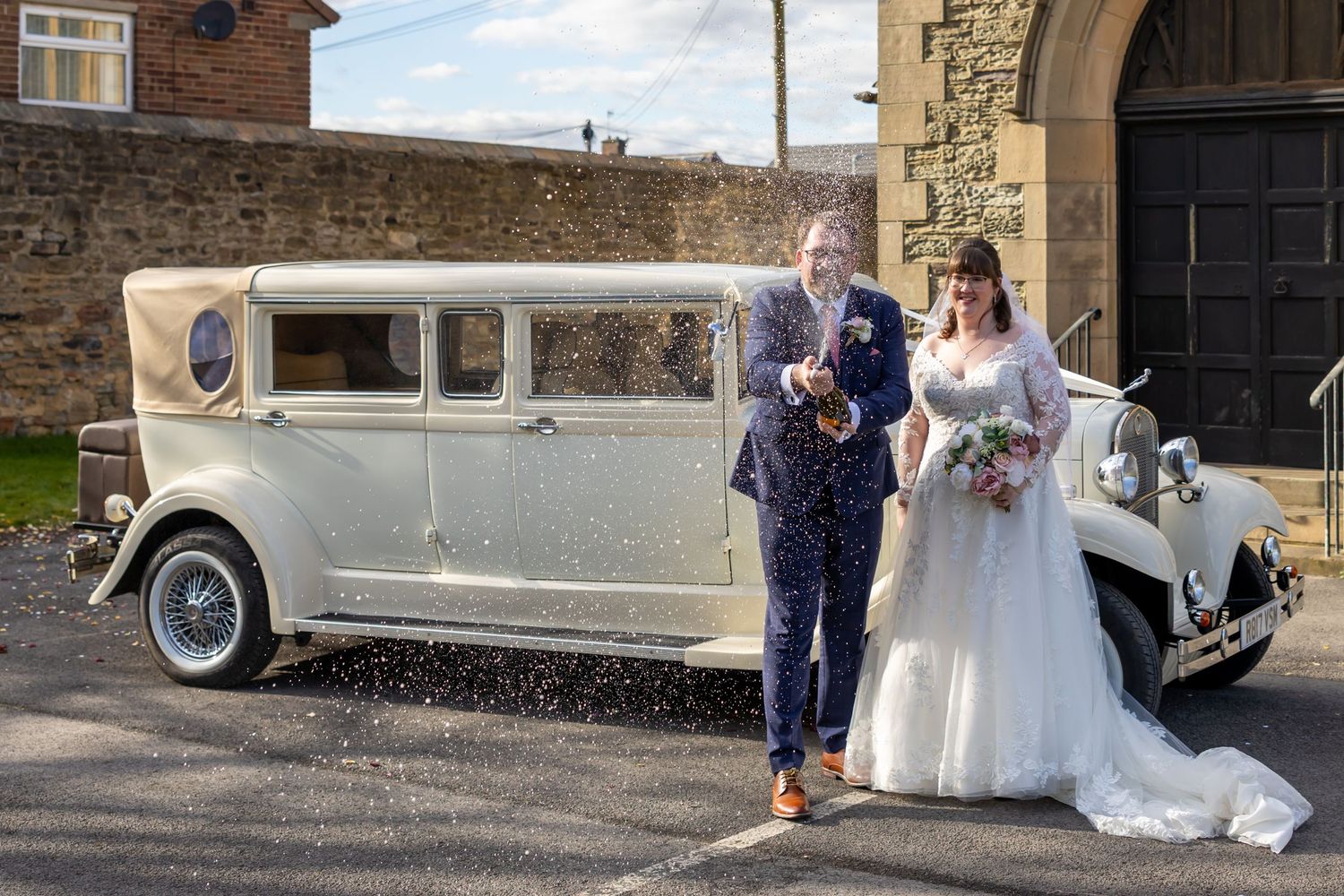 A wedding party poses next to a classic vintage car outside a stone building on a sunny day.