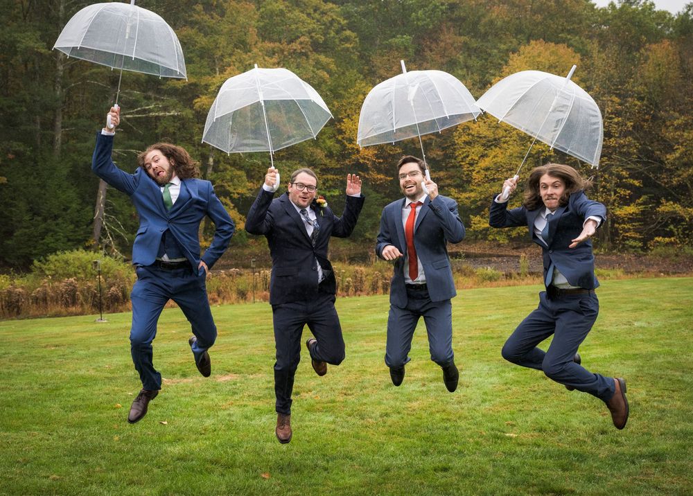 Wedding party jumping in unison on grass while holding umbrellas against autumn foliage background.