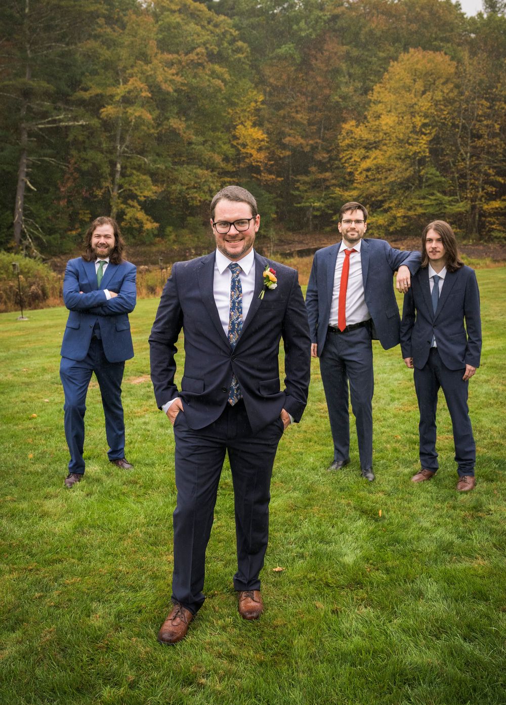 Groomsmen in navy suits pose creatively on a green lawn with autumn trees in the background.