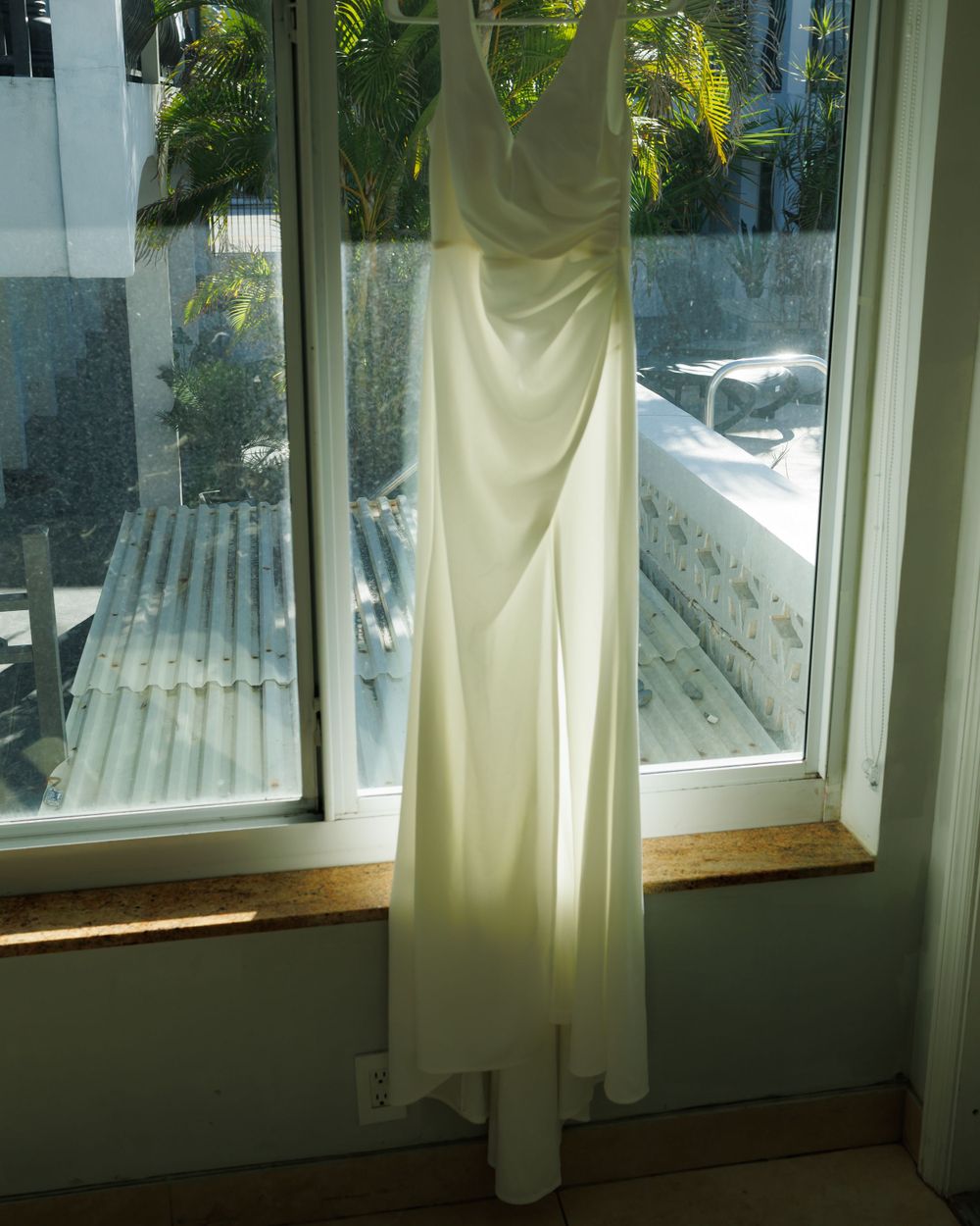 A flowing white wedding dress hangs elegantly in a corner window with tropical palm trees visible outside.