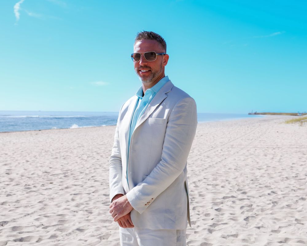Someone in a light gray suit stands on a sunny beach with bright blue sky overhead and sand stretching into the distance.
