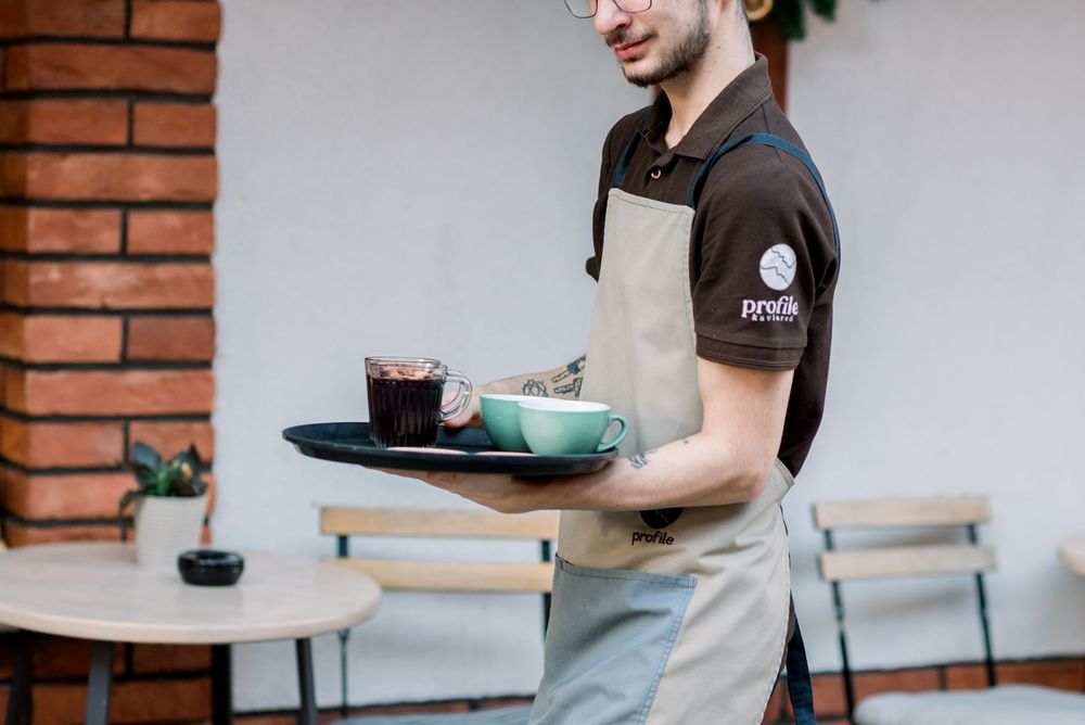 Server in apron carrying drinks and food on tray at outdoor cafe setting.