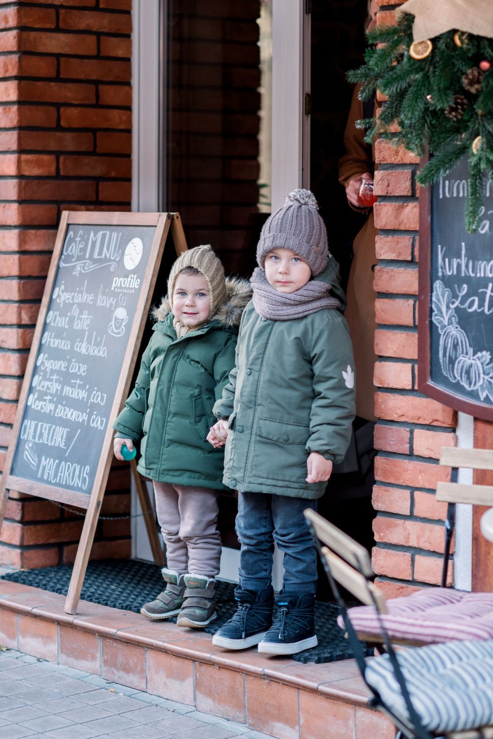 Two children in matching green winter coats stand by a chalkboard menu outside a cafe.