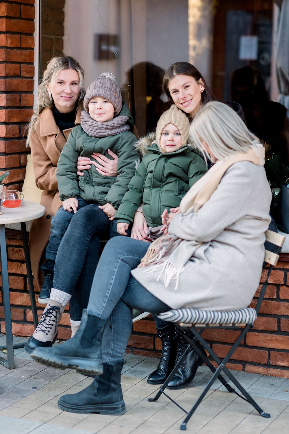 Group gathered at outdoor cafe wearing winter coats and boots against brick wall.