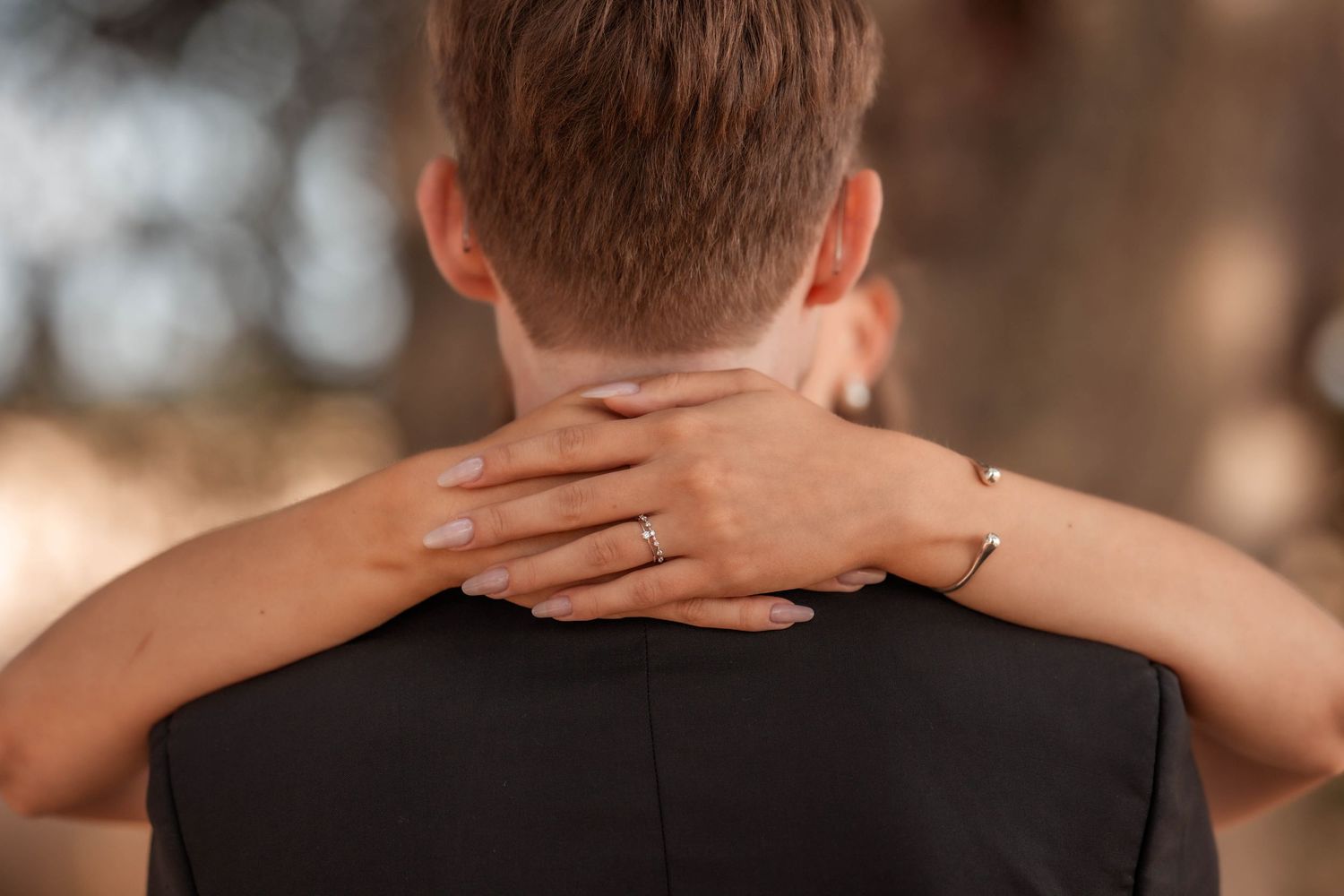 A woman with a bracelet and ring embraces a man from behind, her hand resting on his neck outdoors.
