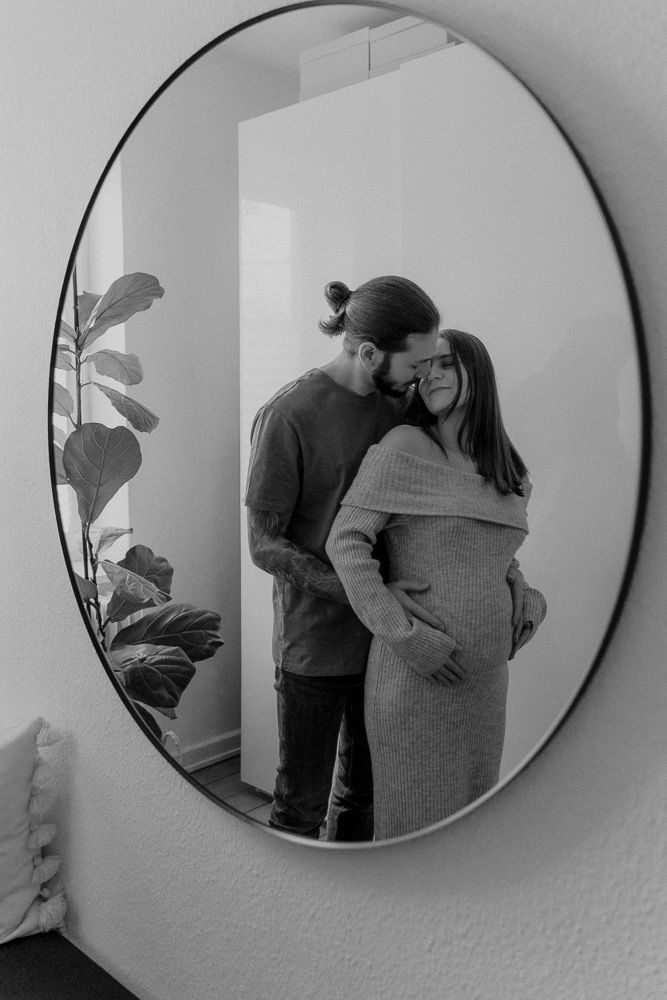 Black and white mirror reflection of an intimate maternity moment showing a couple embracing in a cozy interior setting.