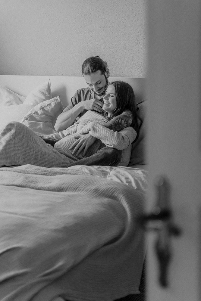 A couple rests peacefully together in bed under cozy blankets in a black and white photograph.
