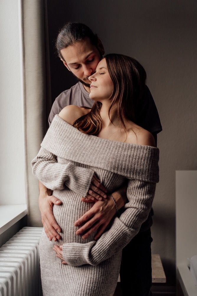 A couple shares an intimate embrace by a window, with one person wearing a cozy off-shoulder sweater.