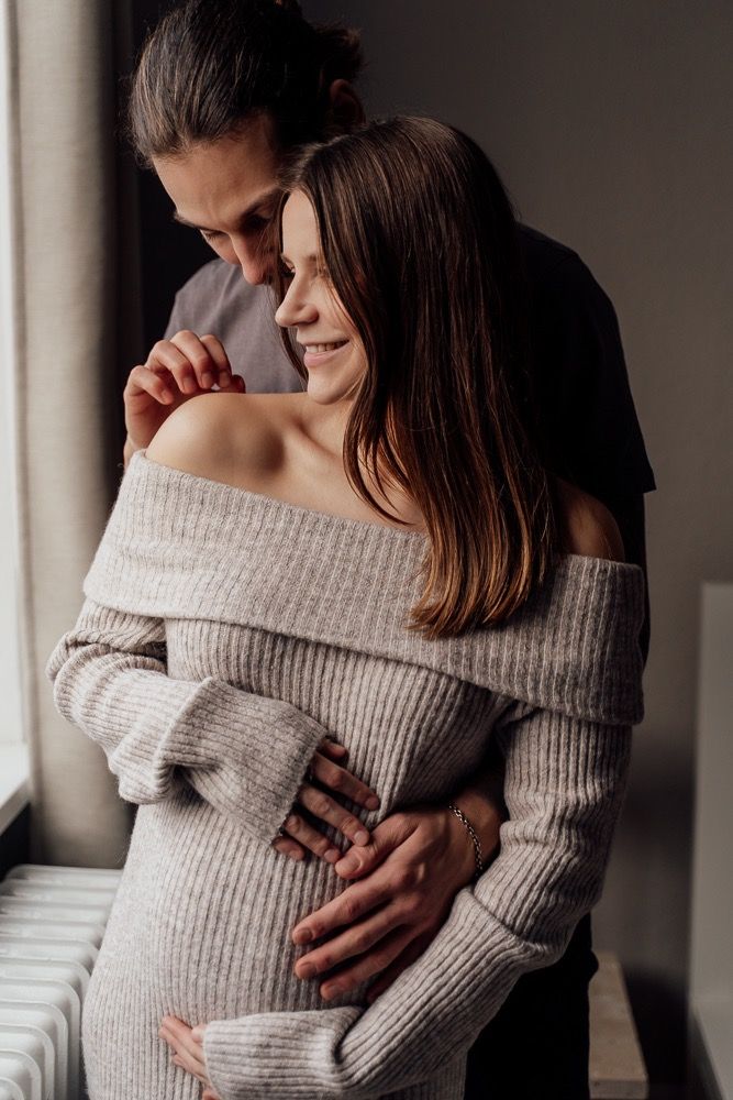 Intimate couple in cozy off-shoulder sweater sharing tender embrace in moody indoor lighting.