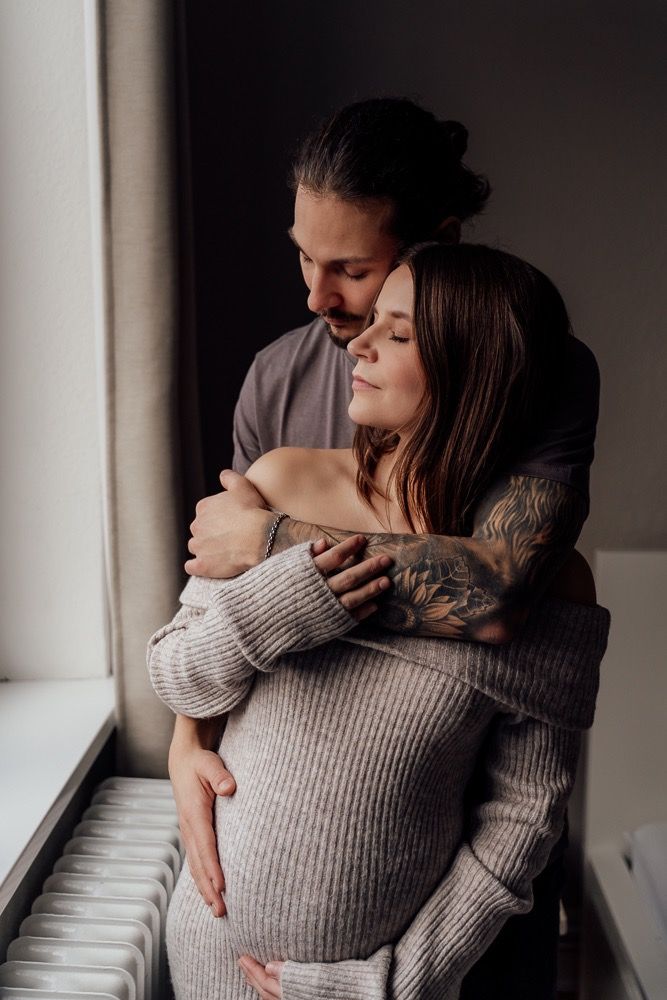 A couple shares an intimate embrace while standing near a window in soft natural light wearing a cozy sweater.