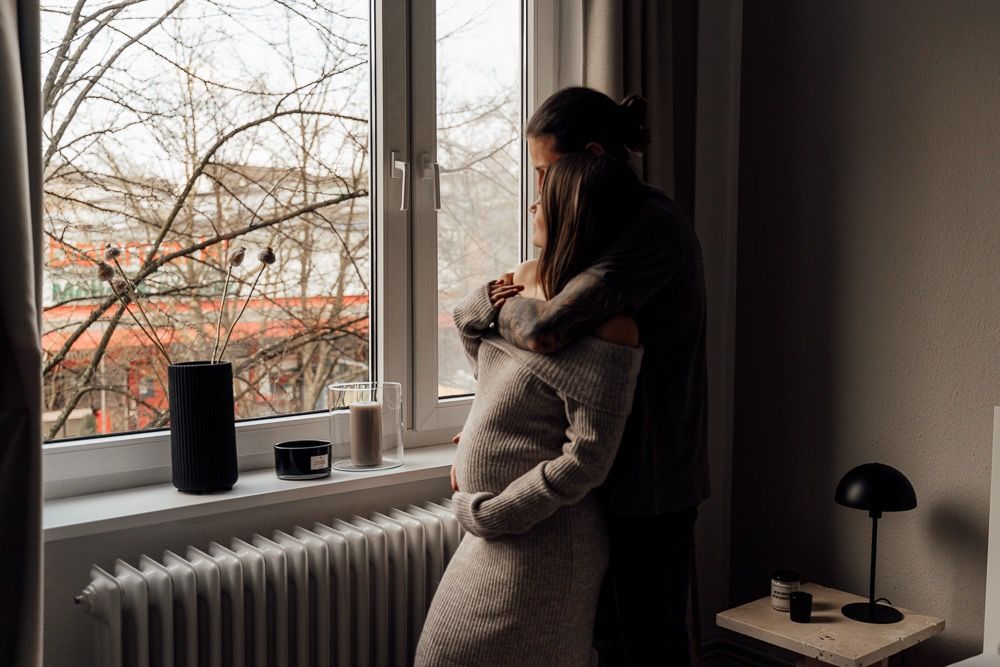 A person in a cozy sweater gazes thoughtfully out a window beside a radiator and desk lamp on a winter day.