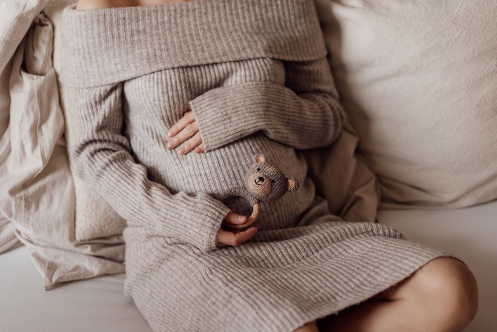 Person wearing cozy gray sweater relaxes on white bedding while holding hot beverage in ceramic mug.
