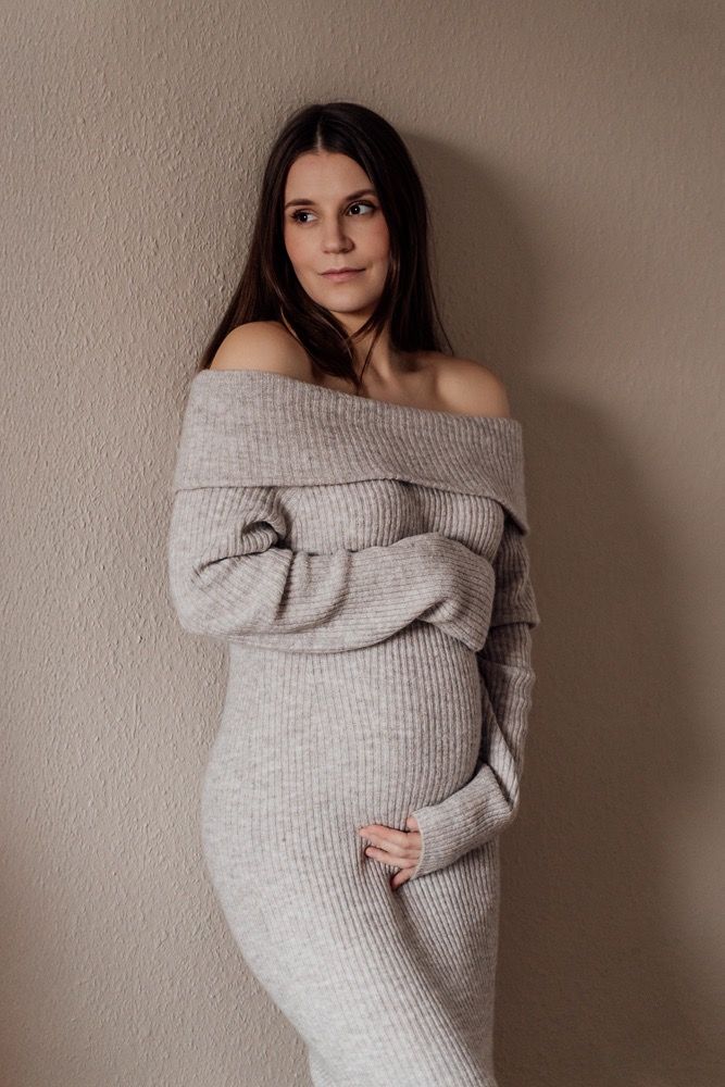 Woman in cozy gray off-shoulder sweater poses against neutral wall in maternity photoshoot series.