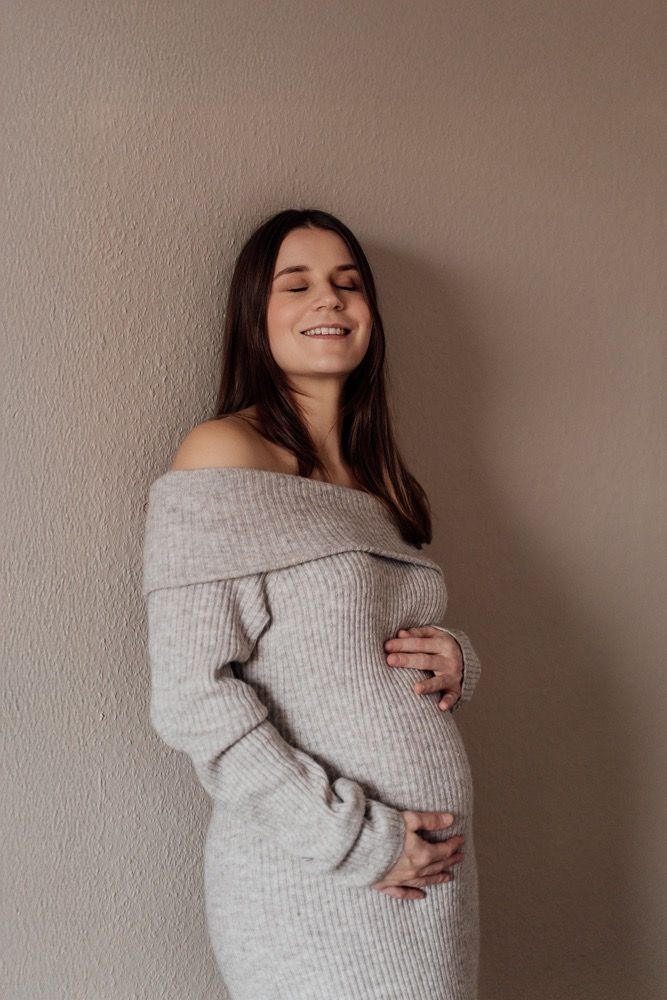 Expectant mother smiles warmly while wearing comfortable gray knit sweater in natural light setting.