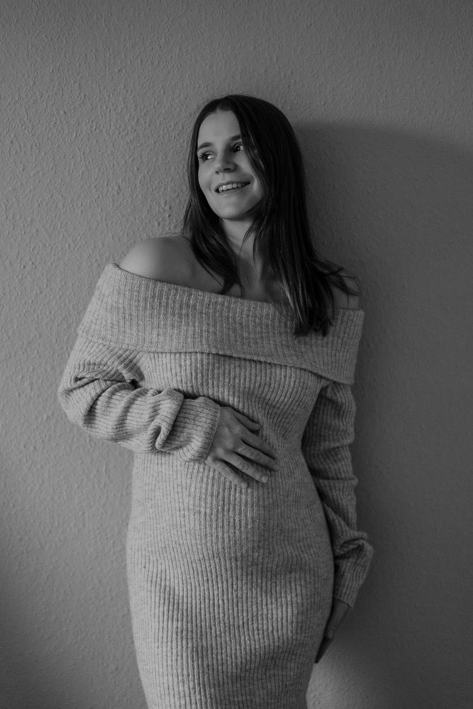 Black and white portrait of someone in a cozy off-shoulder sweater dress leaning against a wall.