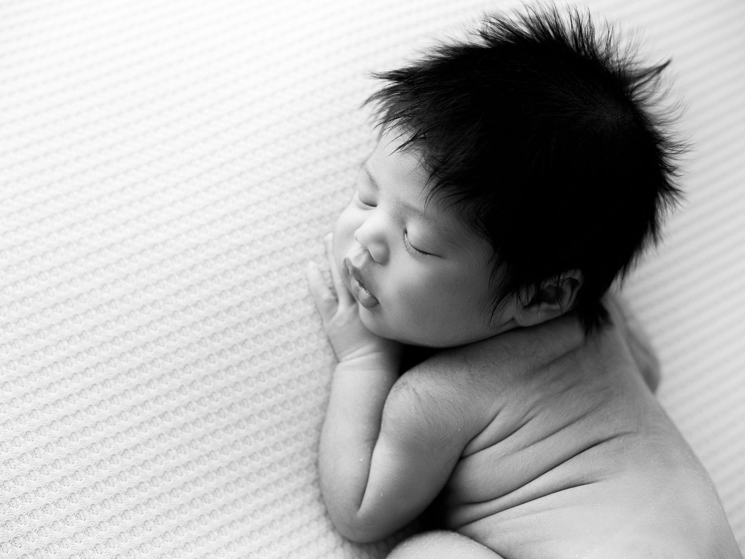 A sleeping baby with spiky black hair lies peacefully on white bedding.
