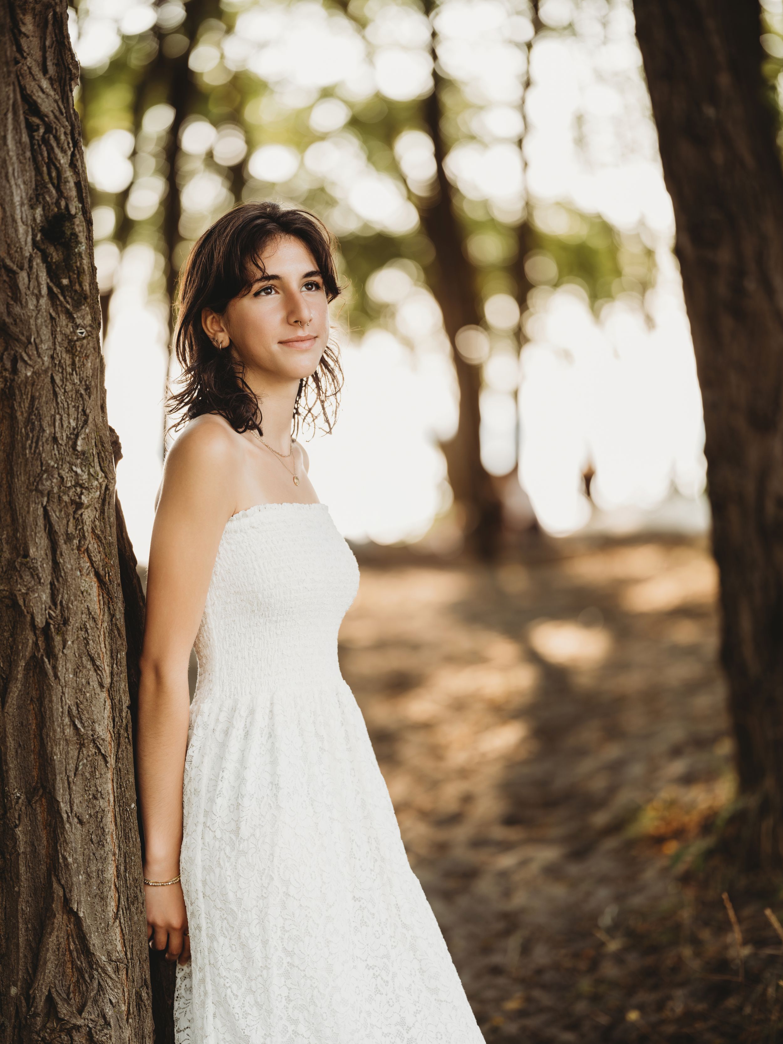 A person in a flowing white lace dress poses thoughtfully among tall pine trees in a forest setting.
