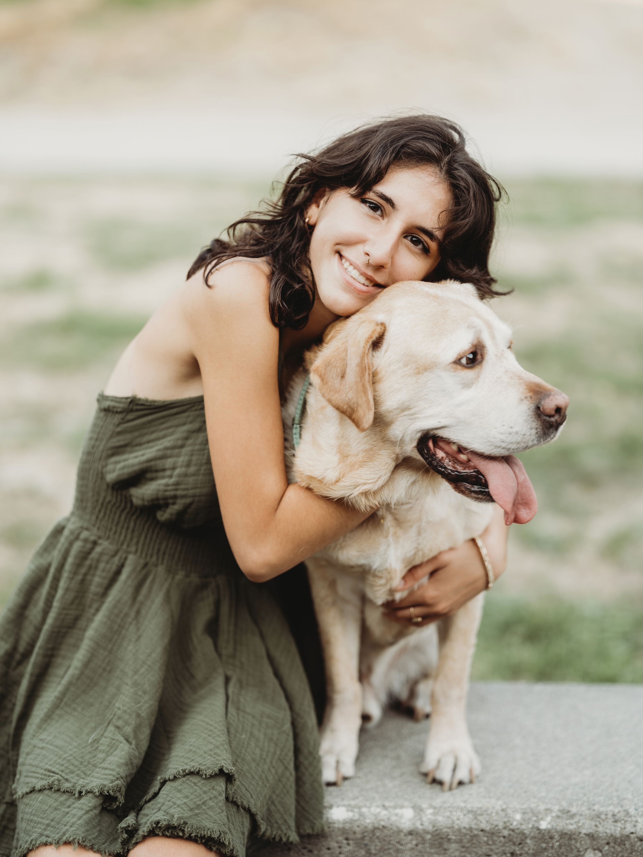 A person in an olive green dress embraces a happy white dog outdoors on a sunny day.