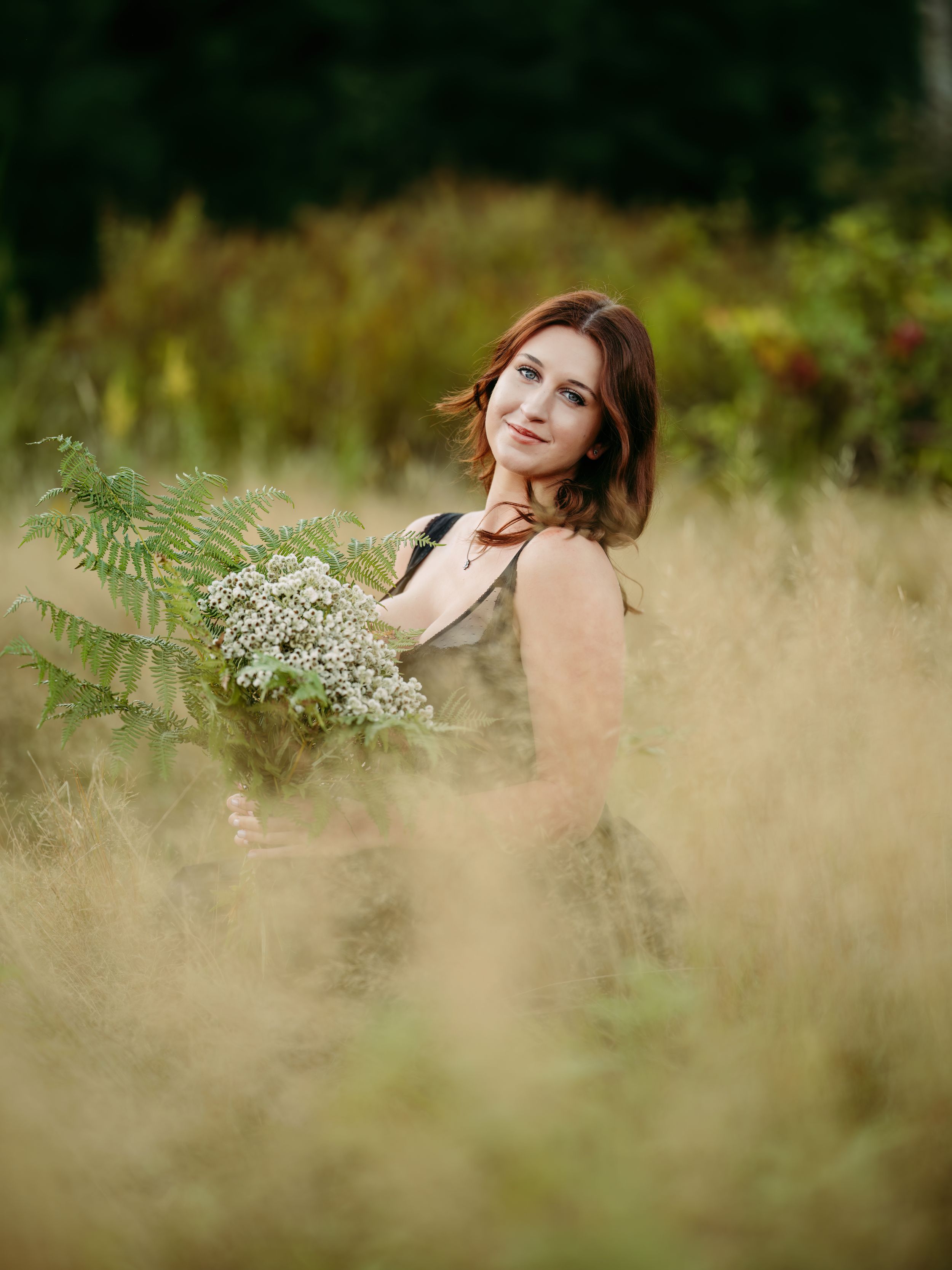 Woman with red hair poses dreamily in a golden meadow with delicate white wildflowers during sunset.