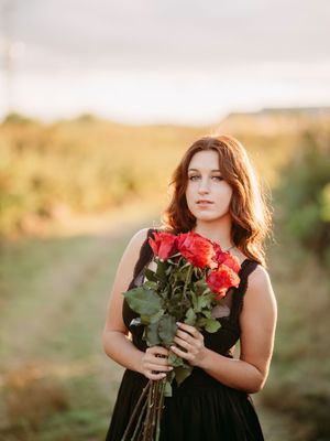 A person in a black dress holds red roses against a golden sunset backdrop in a romantic outdoor setting.