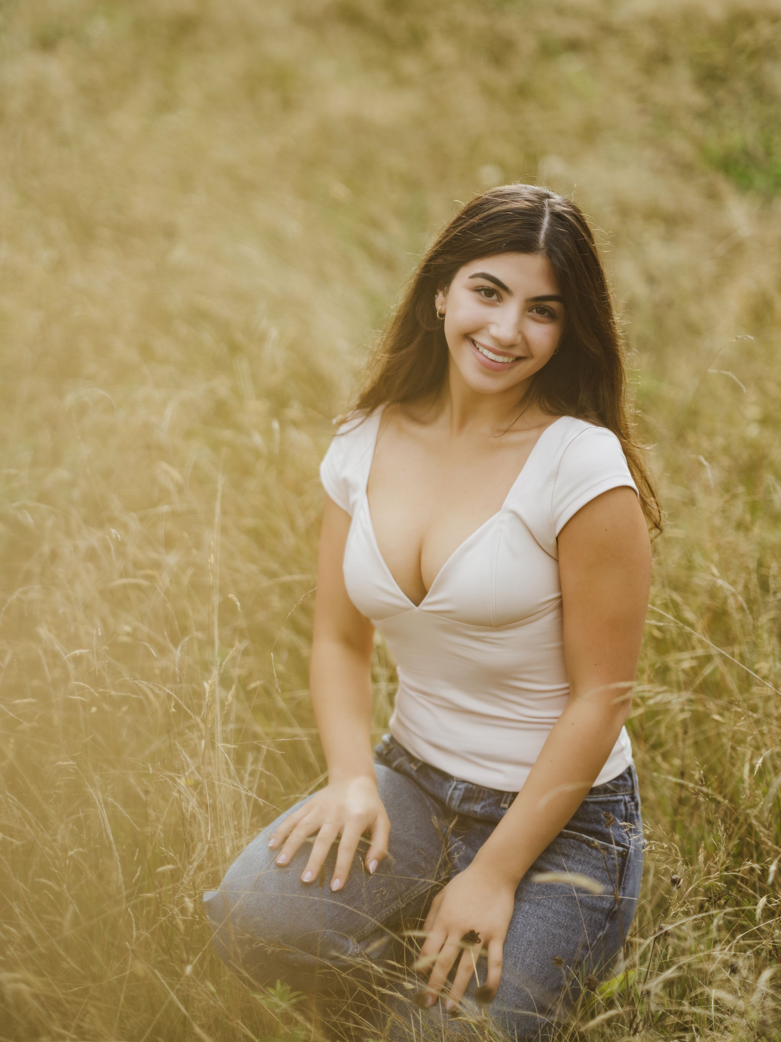 A woman in a white crop top and jeans sits in a golden wheat field during sunset.