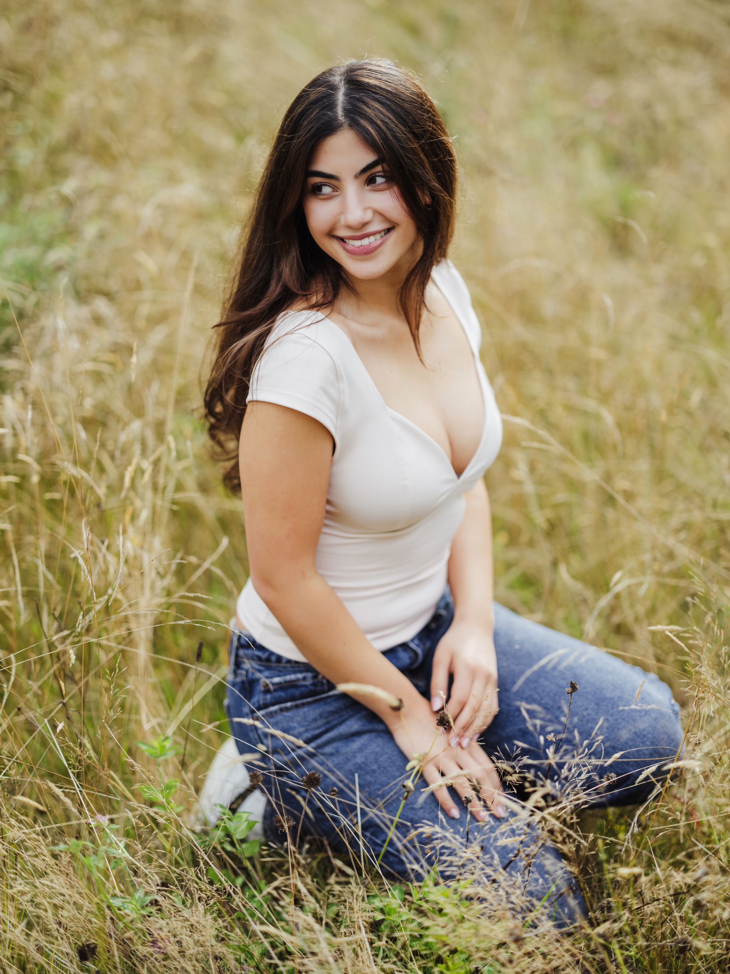 A person in a white top and blue jeans sits casually in a sun-lit field of tall grass during a photo session.