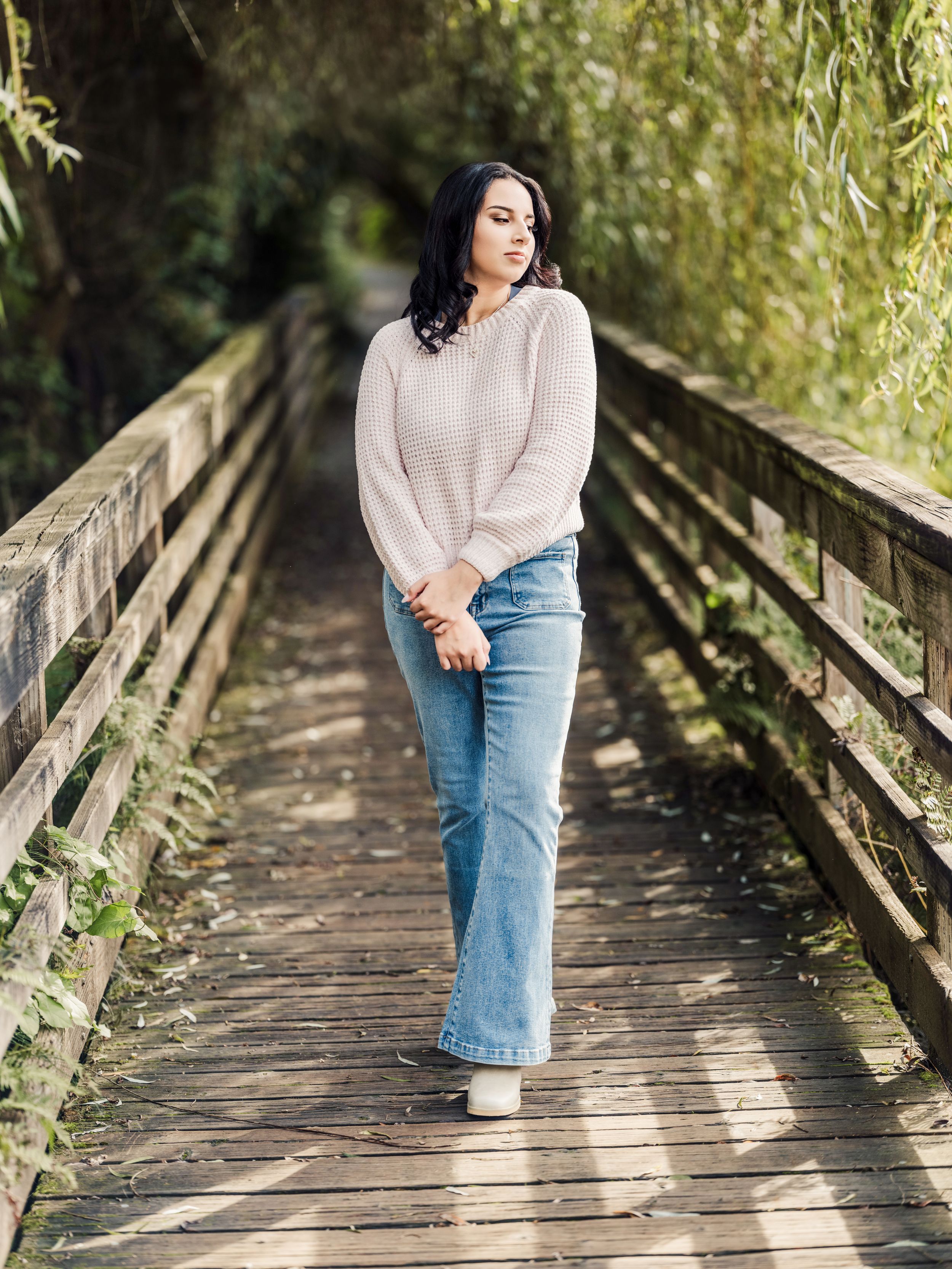 Someone in a cream sweater and jeans walks along a wooden bridge surrounded by willow trees.