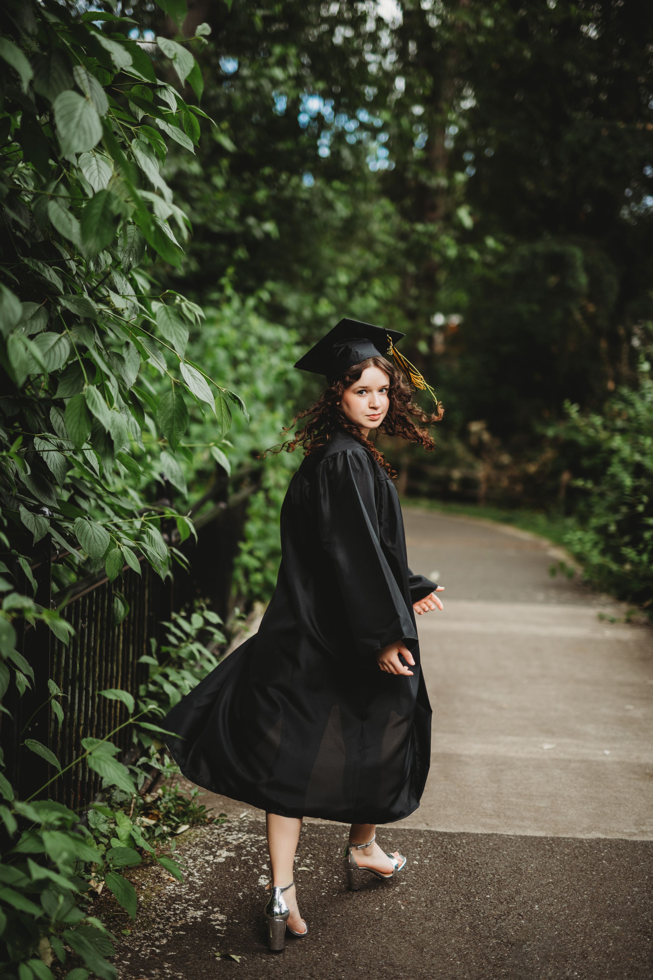 A woman in a flowing black dress poses gracefully on a garden path surrounded by lush green foliage.