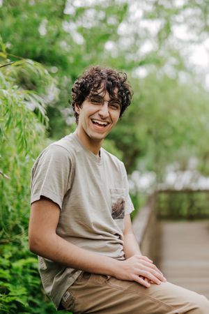 A person in a gray t-shirt sits outdoors smiling and laughing while surrounded by green foliage in a natural setting.