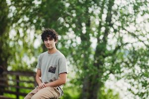 Person sits casually outdoors in a grey t-shirt against a lush green forest background during daytime.