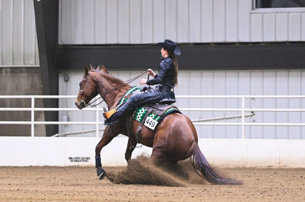A horse and rider perform a sliding stop maneuver in a sandy indoor arena during a reining competition.