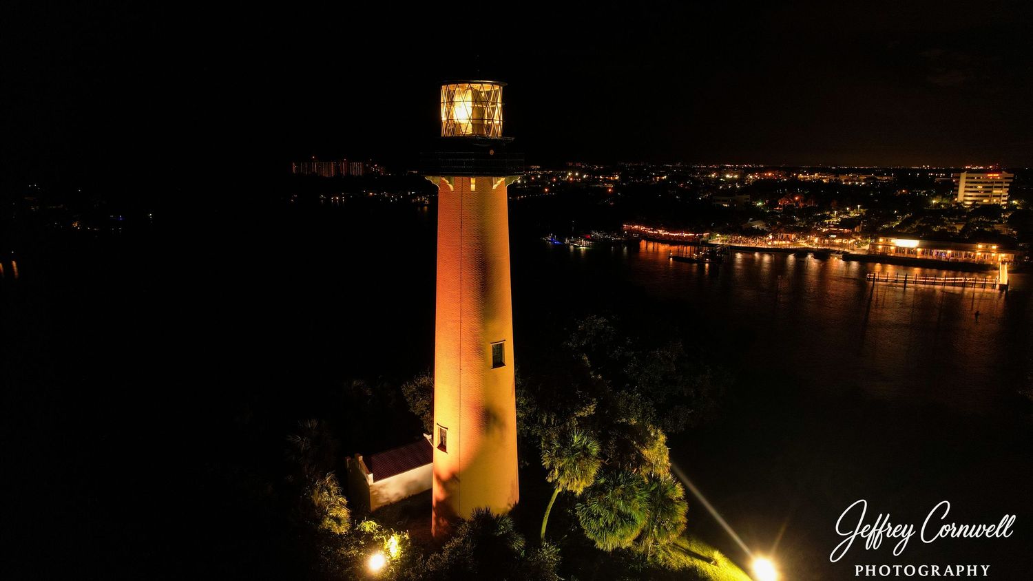 Illuminated lighthouse glows against dark night sky overlooking city lights and waterfront.