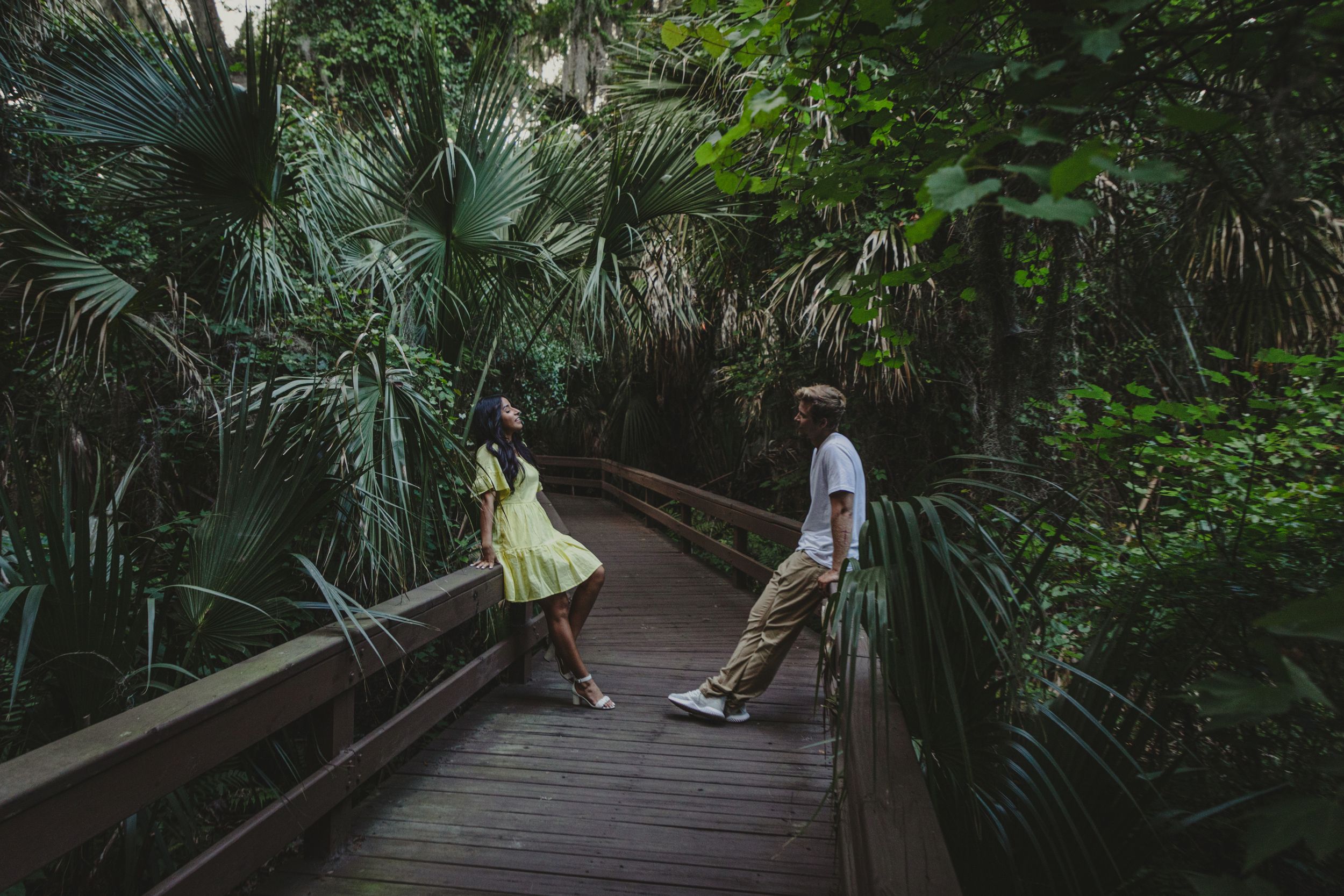 Two people relax on a wooden boardwalk surrounded by lush tropical palms and greenery in a peaceful nature setting.