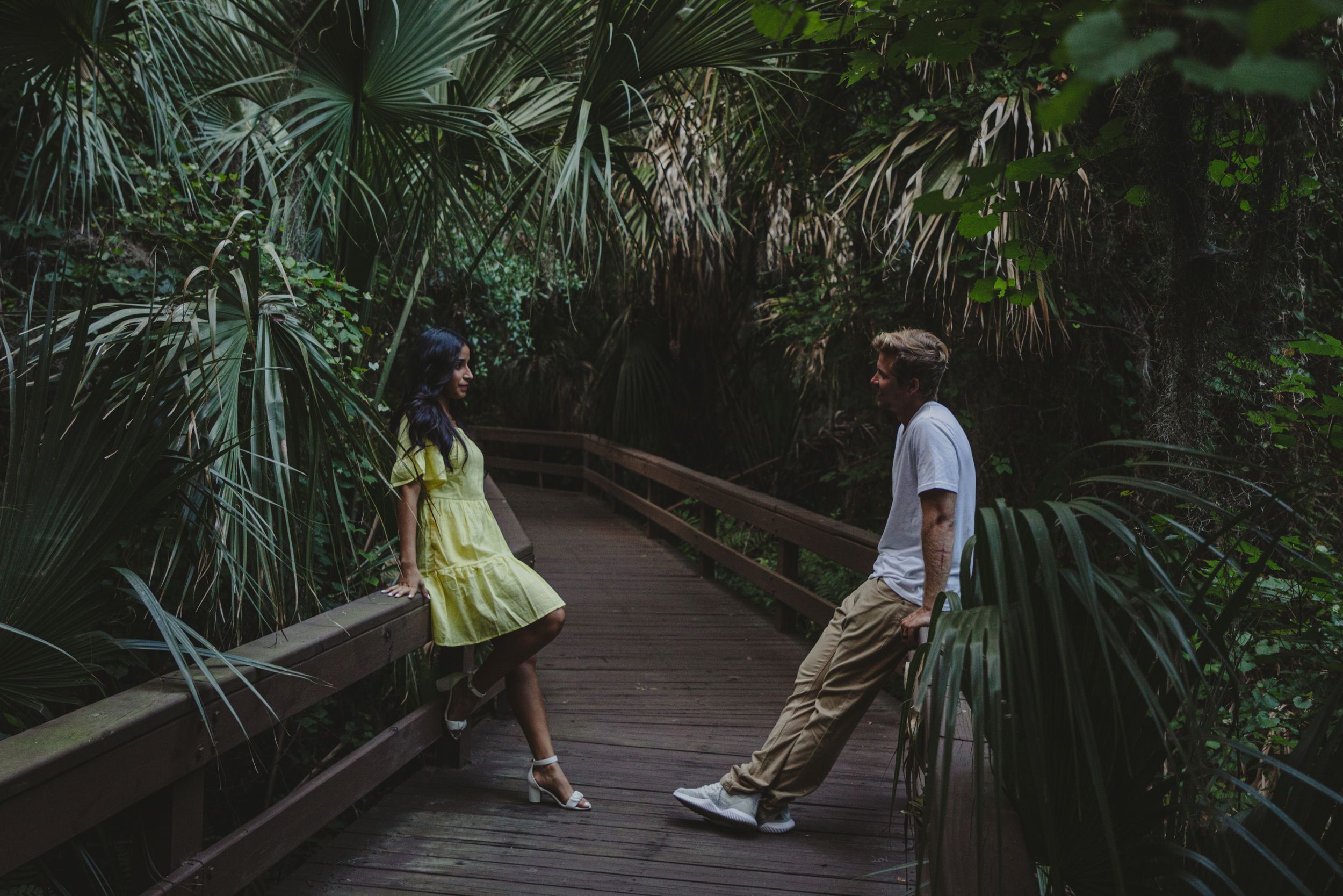 Two people stand apart on a wooden boardwalk surrounded by lush tropical palm fronds in a nature setting.